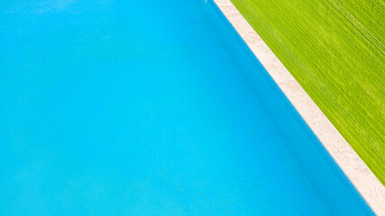 Aerial view of a swimming pool, belonging to a large villa. The pool is empty and no one is swimming. Around the water there is a green grass floor.
