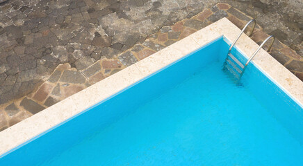 Aerial closeup of a rectangular swimming pool with ladder, belonging to a large villa. The pool is empty and no one is swimming. Around the water there is a stone floor.