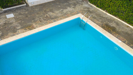 Aerial view of a rectangular swimming pool with ladder, belonging to a large villa. The pool is empty and no one is swimming. Around the water there is a stone floor.