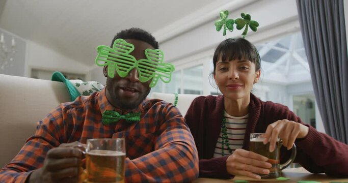 Portrait of happy diverse couple with beer wearing clover shape items and having video call