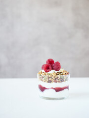 Glass with delicious sweet granola and yogurt decorated with ripe fresh raspberry placed on table against gray wall