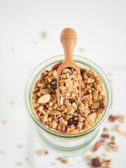High angle of appetizing granola with various nuts and dried berries in glass jar with wooden scoop placed on white table