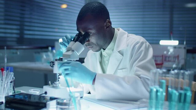 African American scientist in sterile gloves and white coat using microscope and taking notes while doing research in laboratory