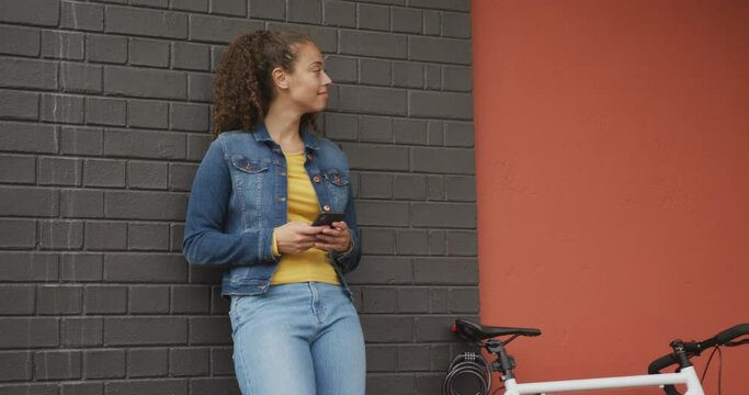 Happy biracial woman in city, next to bike using smartphone