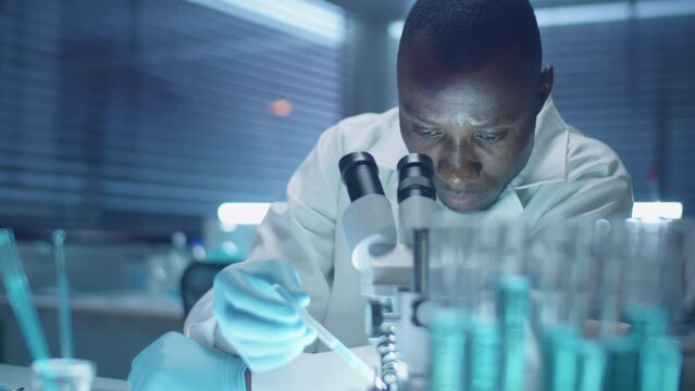 African American scientist in sterile gloves and lab coat examining chemical under microscope while working in laboratory in evening