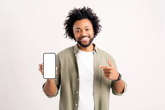 Attractive 30s african-american man pointing finger at smartphone with blank empty screen standing isolated on white, positive curly guy with toothy smile pointing finger at phone device, mock up