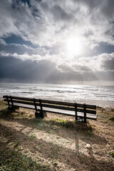 Fototapeta na wymiar Wooden bench at the beach near the ocean at Rivedoux, Re island, France. sun is shining among grey clouds.