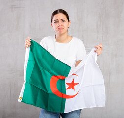 Portrait of disappointed, sad young female holding national flag of Algeria against gray wall