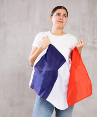Depressed sad young female in casual clothes holding flag of France against gray wall indoors