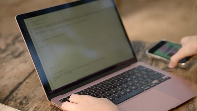 Woman developer working on laptop takes phone with diagrams and checks statistics. Freelancer woman works remotely on computer, typing, coding and checking charts on smartphone. Remote work concept