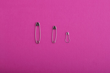 A row of silver safety pins of various sizes and shapes, on a bright pink textured background. Sewing accessories