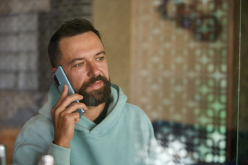 Young businessman talking on his phone indoor through glass. Bearded man in casual clothes using his smartphone.