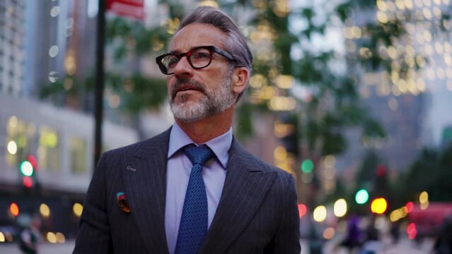 Adult and successful business man in expensive suit, stylish eye glasses walks down street in financial district, he feels confident. 50 years old entrepreneur with gray hair walk in evening city
