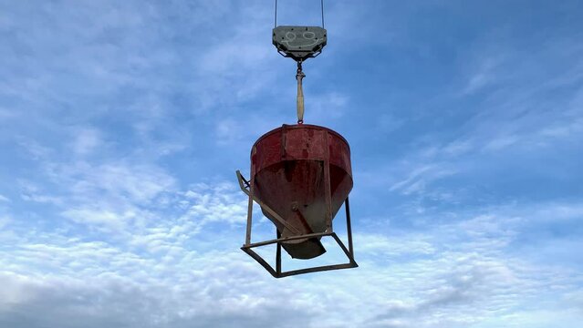 Red bucket with concrete hung from crane in the middle of a blue sky. Bucket of cement holded by a hook with a face painted in it. Steel cement container hanging in the air.