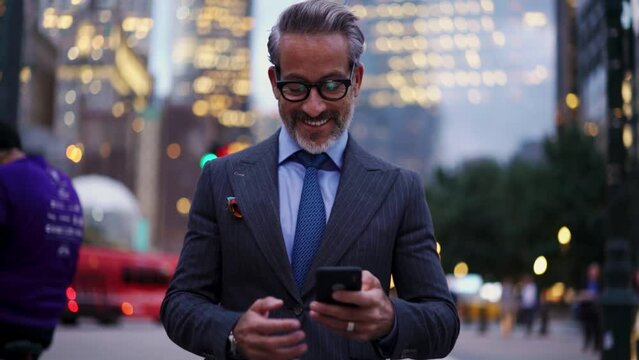 Adult And Happy Successful Business Man In Expensive Suit, Stylish Eye Glasses Walks Down Street Of New York, He Feels Confident. 50 Years Old Male Use Mobile Phone Device While Smiling And Rejoices