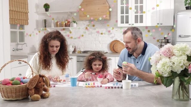 Charming family having good time together. Portrait of a beautiful, caucasian couple helping their cute daughter to paint traditional Easter eggs on holiday. High quality 4k footage