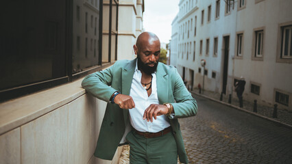 A sophisticated bald black man wearing a pastel green suit and a white shirt, accessorized with necklaces and checks the time on his wristwatch while standing in the middle of a cobblestone street - obrazy, fototapety, plakaty