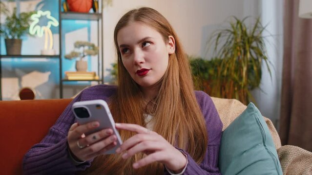 Woman sitting on sofa uses mobile phone smiles at home living room apartment. Portrait of young girl texting share messages content on smartphone social media applications online, watching relax movie