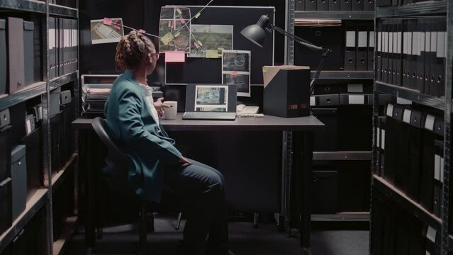 African american woman examining evidence map to find suspect, using detective board on office wall. Young police officer analyzing research information for investigation, archive room.