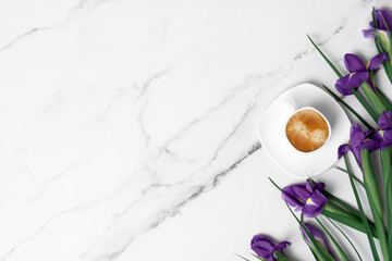 Cup of hot morning coffee and violet irises on marble background. Flat lay with copy space for...