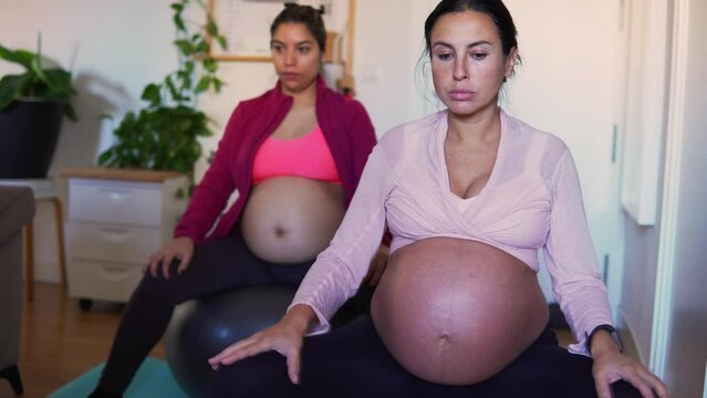 Multiethnic pregnant women doing yoga at home using pilates balls. Two pregnant women bellies while relaxing with circular movements