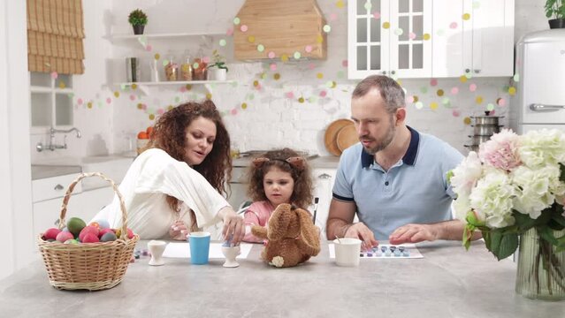 Caucasian parents playing with their cute daughter in home kitchen with beautiful decorations. Portrait of adorable family painting Easter eggs. High quality 4k footage