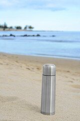 Metallic thermos with hot drink on sandy beach near sea, space for text