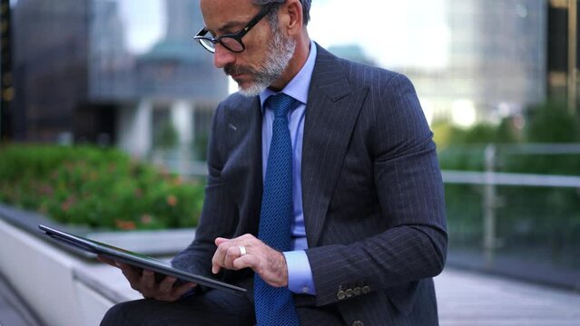 Successful Caucasian businessman in stylish eye glasses planning work on touch pad while touching screen. Pensive mature entrepreneur in suit sitting outdoors with modern tablet develops strategy idea
