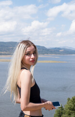 Beautiful young woman with long blond hair in a black top and shorts standing in front of a lake and holding a mobile phone