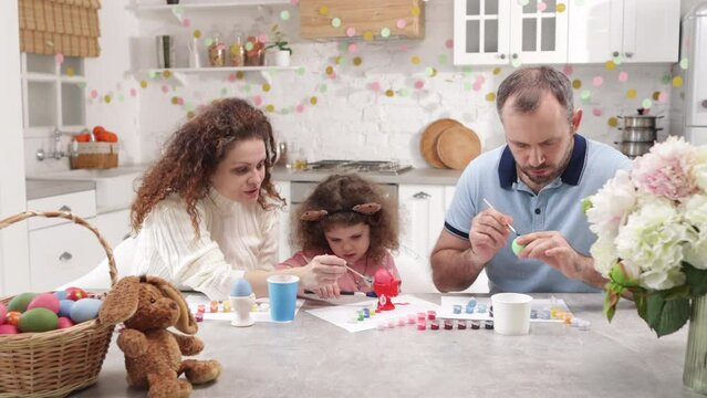 Laughing, happy mother and father helping their girl to paint Easter symbol. Portrait of happy family with adorable daughter painting Easter eggs in the kitchen. High quality 4k footage