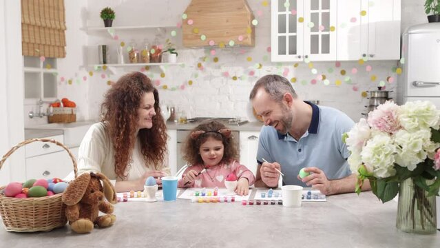 Portrait of a beautiful, caucasian couple helping their cute daughter to paint traditional Easter eggs on holiday. Mom and dad laughing and having fun in a charming home. High quality 4k footage