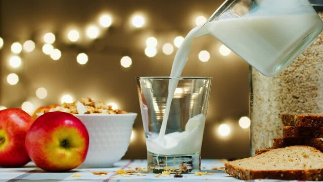 Pouring milk into a glass from a jug against the background of bright light bulbs. Fresh milk in a jug is poured into a glass, slow motion. healthy eating, soya rice coconut skim milk, nutrition