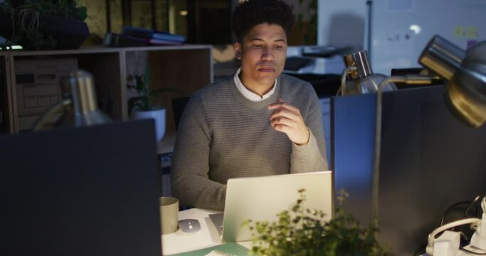 Video of focused biracial man using laptop, working late in office