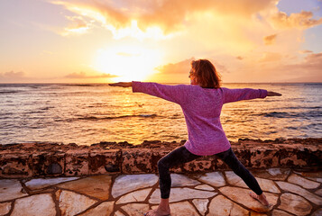 Fototapeta premium Active Senior Woman practicing yoga postures outside by the ocean during sunset