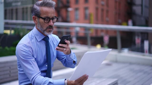 Focused mature businessman in blue shirt sits on street and studies paper documents, taking photo on mobile phone. 50 years old financial expert reading report while sitting outdoors in downtown