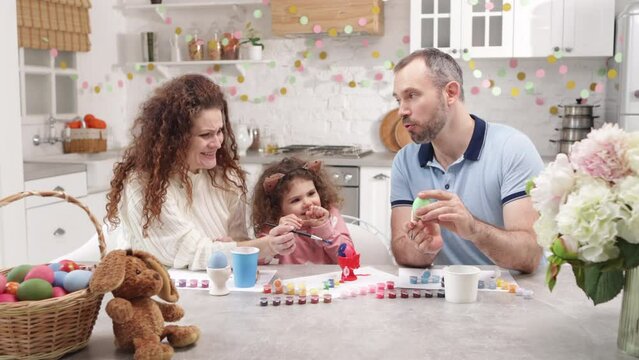 Caucasian parents in their 40s having fun with their cute baby girl. Portrait of a family painting colorful Easter eggs for a festive season. High quality 4k footage