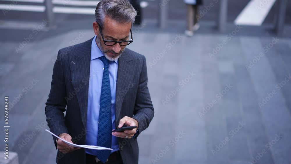 Poster successful businessman standing on city street with paper documents and typing text message in smart