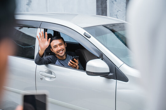 male driver in a car holding a cell phone and waving as he meets customers ordering online