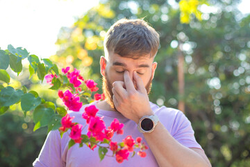 Portrait of handsome young allergic man is suffering from pollen allergy or cold on natural flowers, flowering tree background at spring or sunny summer day, sneezes, blowing his runny nose rubs eyes - obrazy, fototapety, plakaty