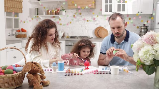 Portrait of happy family with adorable daughter painting Easter eggs in the kitchen. Caucasian parents in their 40s having fun with their cute baby girl. High quality 4k footage