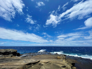 Ocean waves crashing in water on rock formations with blue skies on a sunny day with some clouds in Oahu Hawaii