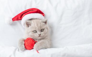 Funny kitten wearing red santa's hat hugs ball of wool under white warm blanket. Top down view....