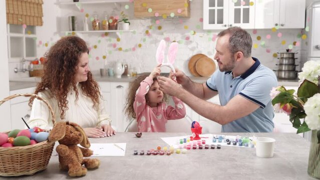 Mom and dad wearing cute bunny ears with their little daughter. Portrait of a charming family preparing for Easter holidays in a beautifully decorated domestic kitchen. High quality 4k footage