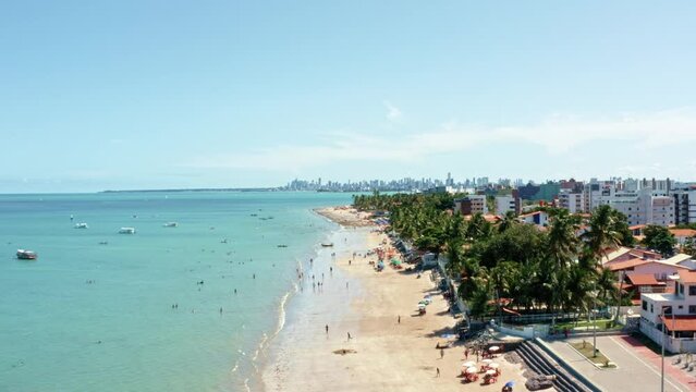 Rising aerial wide shot of Bessa beach in the tropical capital Joao Pessoa, Brazil  in the state of Paraiba with Brazilians and tourists enjoying the ocean and fishing boats at shore on a summer day.