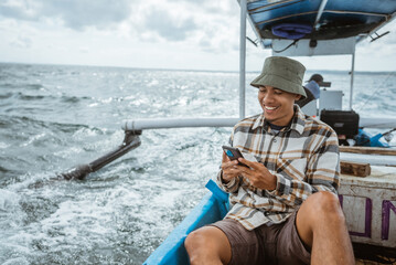 Asian angler takes a break sitting while using his cell phone on a fishing boat