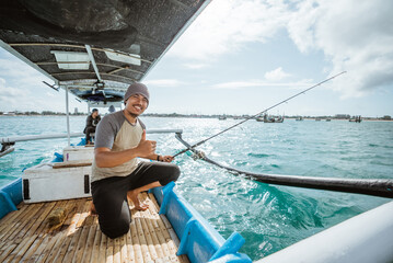 Asian angler with thumbs up while holding a fishing rod over a small fishing boat