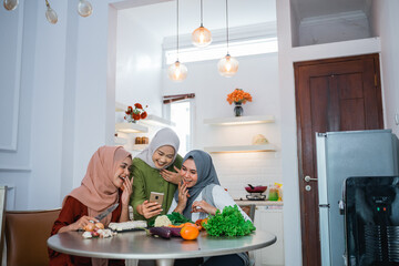 happy young asian woman with hijab cooking in the kitchen while looking at internet on mobile phone for menu today