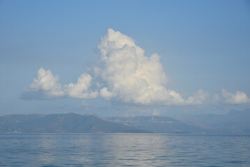 Mediterranean Sea shore view, soft reflection of white cloud in the blue water. Wind turbines on the top of hazy distant mountains seem tiny. Carefree feel of summer day. Beach near Athens, Greece.  