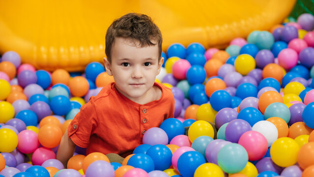 Portrait of a happy little boy jumping merrily into a dry pool with colorful balloons. A toddler boy is playing and having a good time at the children's entertainment center.