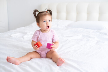 funny baby girl in pink bodysuit playing with children wooden cubes toys on bed